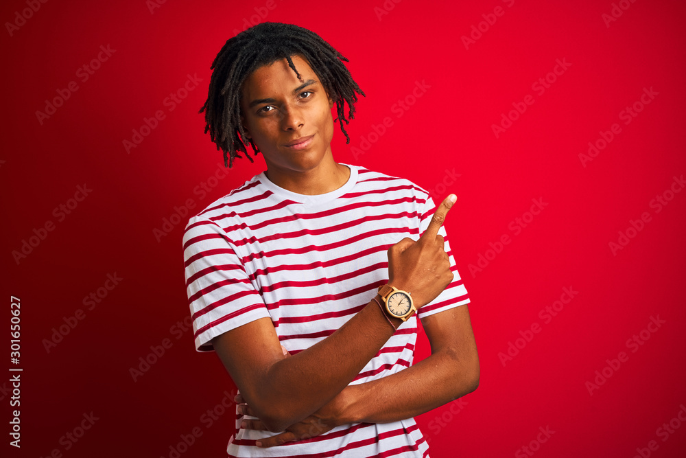 Young afro man with dreadlocks wearing striped t-shirt standing over isolated red background Pointing with hand finger to the side showing advertisement, serious and calm face Young afro man with dreadlocks wearing striped t-shirt standing over isolated red background Pointing with hand finger to the side showing advertisement, serious and calm face