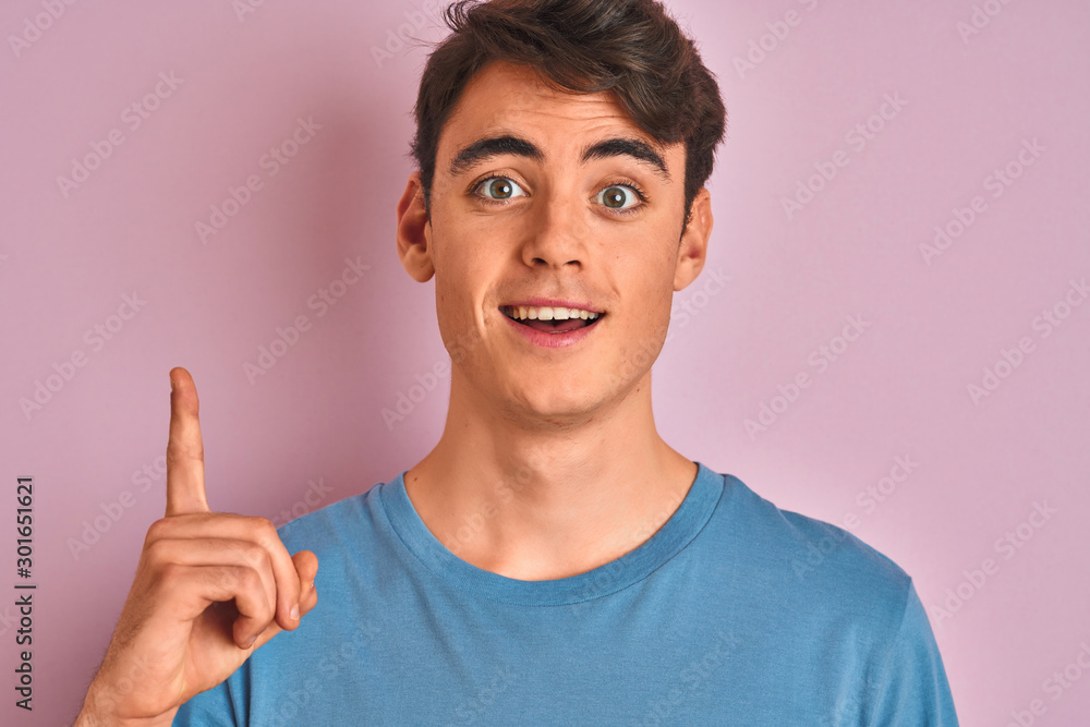 Teenager boy wearing casual t-shirt standing over blue isolated background surprised with an idea or question pointing finger with happy face, number one Teenager boy wearing casual t-shirt standing over blue isolated background surprised with an idea or question pointing finger with happy face, number one