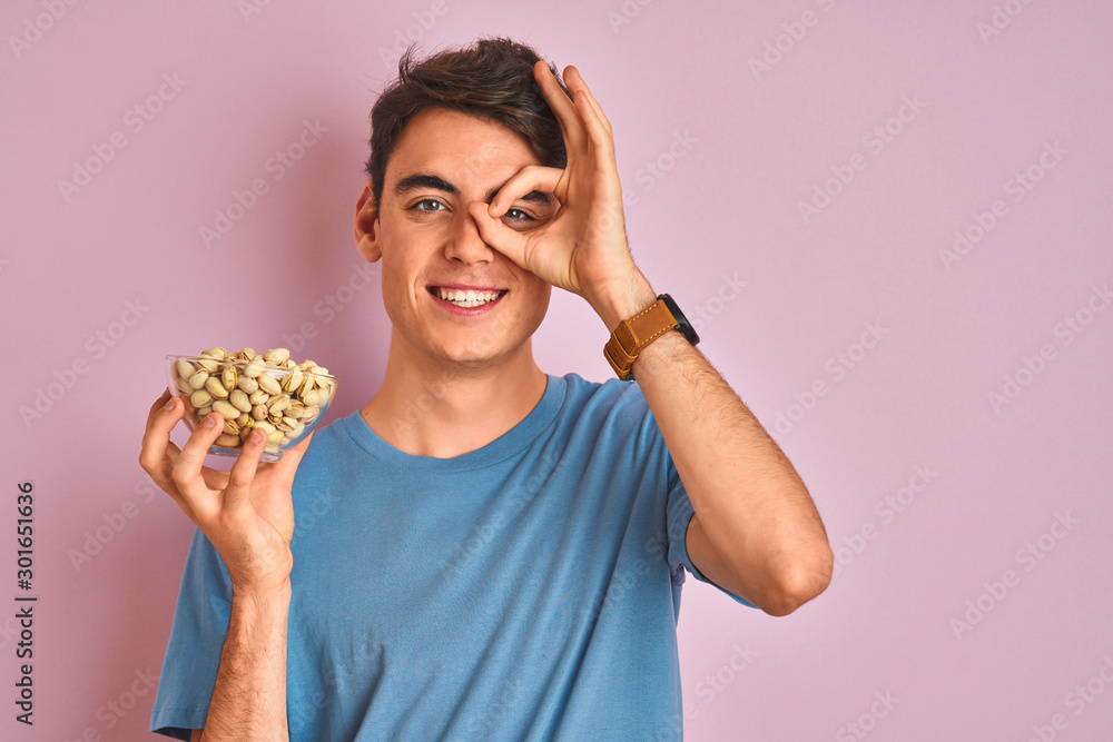 Teenager boy holding a bunch of pistachios over isolated pink background with happy face smiling doing ok sign with hand on eye looking through fingers Teenager boy holding a bunch of pistachios over isolated pink background with happy face smiling doing ok sign with hand on eye looking through fingers