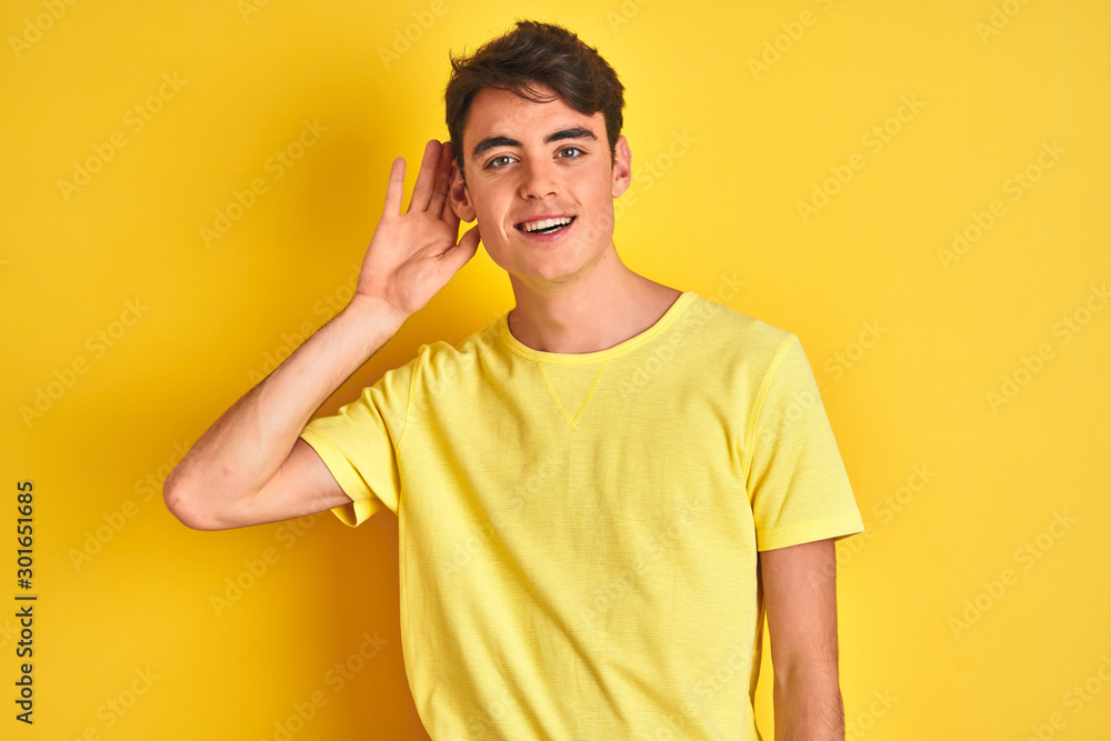 Teenager boy wearing yellow t-shirt over isolated background smiling with hand over ear listening an hearing to rumor or gossip. Deafness concept. Teenager boy wearing yellow t-shirt over isolated background smiling with hand over ear listening an hearing to rumor or gossip. Deafness concept.
