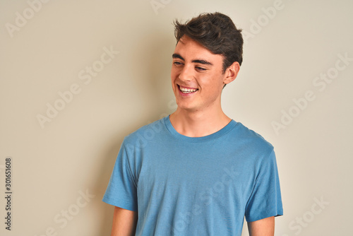 Teenager boy wearing casual t-shirt standing over isolated background looking away to side with smile on face, natural expression. Laughing confident.