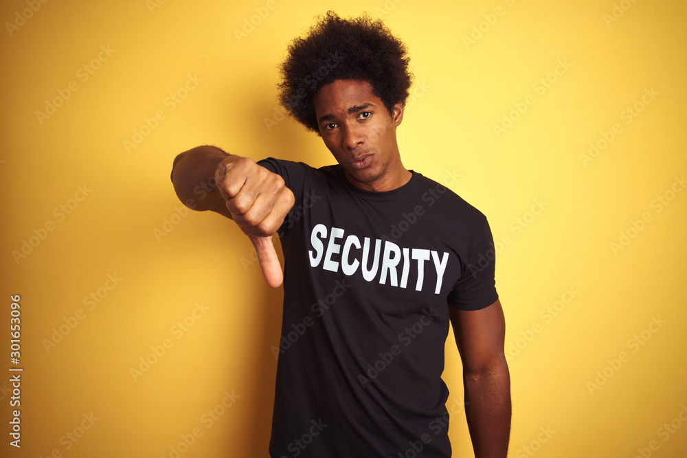American safeguard man with afro hair wearing security uniform over isolated yellow background looking unhappy and angry showing rejection and negative with thumbs down gesture. Bad expression. American safeguard man with afro hair wearing security uniform over isolated yellow background looking unhappy and angry showing rejection and negative with thumbs down gesture. Bad expression.