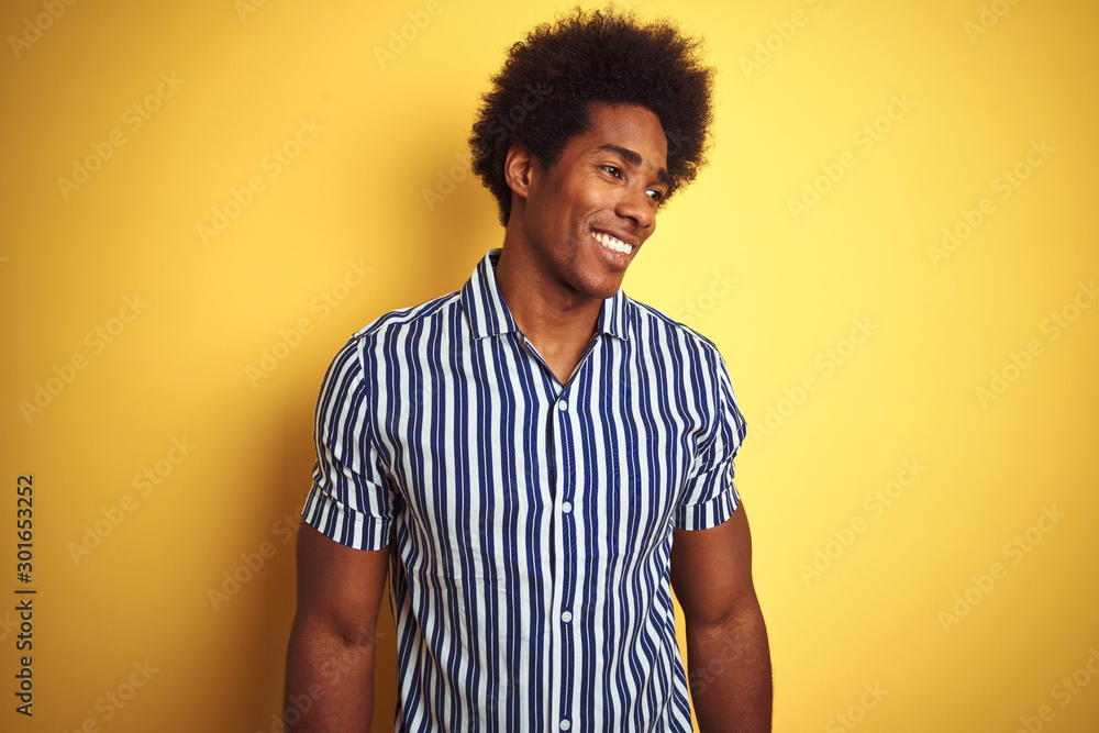 American man with afro hair wearing striped shirt standing over isolated yellow background looking away to side with smile on face, natural expression. Laughing confident. American man with afro hair wearing striped shirt standing over isolated yellow background looking away to side with smile on face, natural expression. Laughing confident.