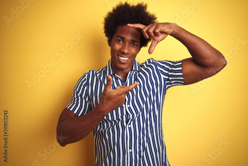 American man with afro hair wearing striped shirt standing over isolated yellow background smiling making frame with hands and fingers with happy face. Creativity and photography concept.