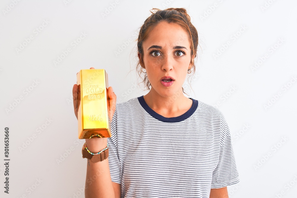 Young beautiful redhead woman holding gold ingot over isolated background scared in shock with a surprise face, afraid and excited with fear expression
