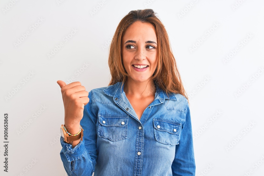 Young redhead woman wearing denim jacket standing over isolated background smiling with happy face looking and pointing to the side with thumb up. Young redhead woman wearing denim jacket standing over isolated background smiling with happy face looking and pointing to the side with thumb up.
