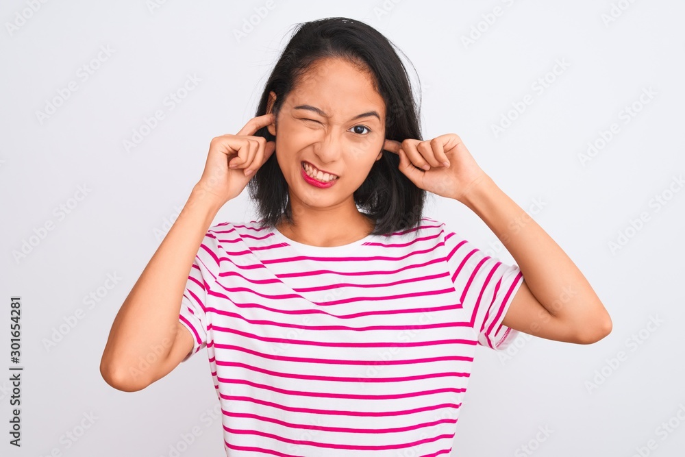 Young chinese woman wearing striped t-shirt standing over isolated white background covering ears with fingers with annoyed expression for the noise of loud music. Deaf concept. Young chinese woman wearing striped t-shirt standing over isolated white background covering ears with fingers with annoyed expression for the noise of loud music. Deaf concept.