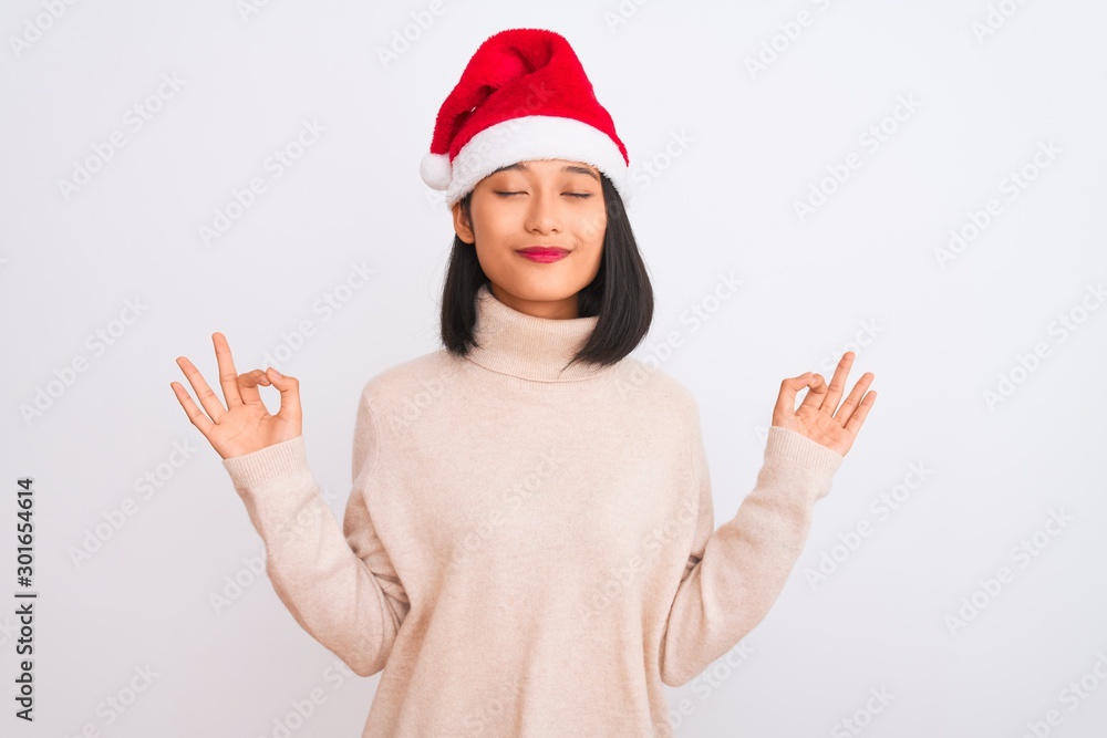 Young beautiful chinese woman wearing Christmas Santa hat over isolated white background relax and smiling with eyes closed doing meditation gesture with fingers. Yoga concept. Young beautiful chinese woman wearing Christmas Santa hat over isolated white background relax and smiling with eyes closed doing meditation gesture with fingers. Yoga concept.