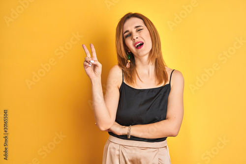 Redhead caucasian business woman standing over yellow isolated background smiling with happy face winking at the camera doing victory sign. Number two. © Krakenimages.com
