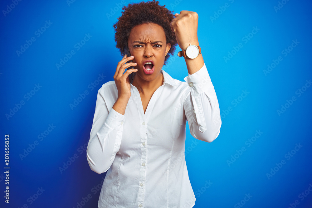 African american woman talking on the smartphone over blue isolated background annoyed and frustrated shouting with anger, crazy and yelling with raised hand, anger concept African american woman talking on the smartphone over blue isolated background annoyed and frustrated shouting with anger, crazy and yelling with raised hand, anger concept