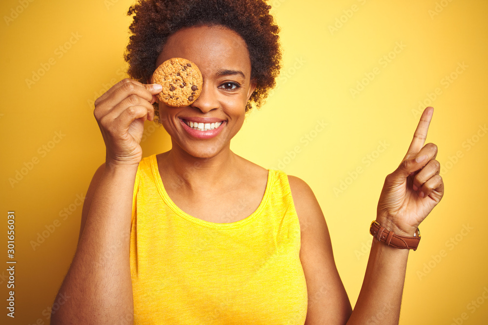 African american woman holding chocolate chips cookie over yellow background very happy pointing with hand and finger to the side African american woman holding chocolate chips cookie over yellow background very happy pointing with hand and finger to the side