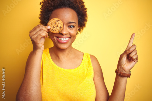 African american woman holding chocolate chips cookie over yellow background very happy pointing with hand and finger to the side