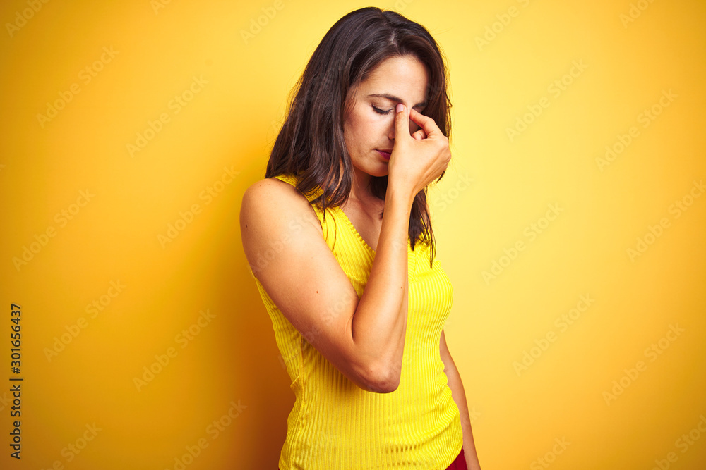Young beautiful woman wearing t-shirt standing over yellow isolated background tired rubbing nose and eyes feeling fatigue and headache. Stress and frustration concept. Young beautiful woman wearing t-shirt standing over yellow isolated background tired rubbing nose and eyes feeling fatigue and headache. Stress and frustration concept.