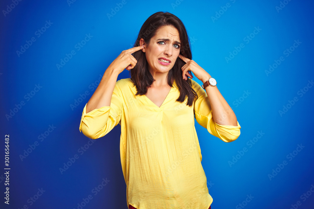 Young beautiful woman wearing yellow t-shirt standing over blue isolated background covering ears with fingers with annoyed expression for the noise of loud music. Deaf concept. Young beautiful woman wearing yellow t-shirt standing over blue isolated background covering ears with fingers with annoyed expression for the noise of loud music. Deaf concept.