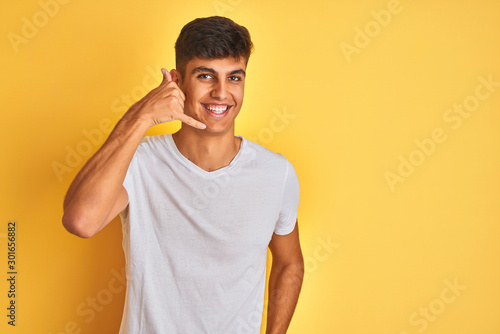 Young indian man wearing white t-shirt standing over isolated yellow background smiling doing phone gesture with hand and fingers like talking on the telephone. Communicating concepts.