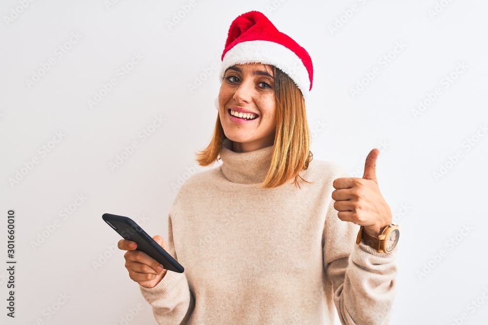 Beautiful redhead woman wearing christmas hat and using smartphone happy with big smile doing ok sign, thumb up with fingers, excellent sign Beautiful redhead woman wearing christmas hat and using smartphone happy with big smile doing ok sign, thumb up with fingers, excellent sign