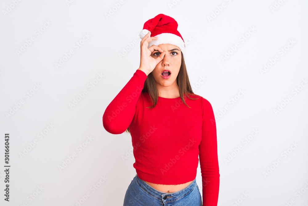Young beautiful girl wearing Christmas Santa hat standing over isolated white background doing ok gesture shocked with surprised face, eye looking through fingers. Unbelieving expression. Young beautiful girl wearing Christmas Santa hat standing over isolated white background doing ok gesture shocked with surprised face, eye looking through fingers. Unbelieving expression.