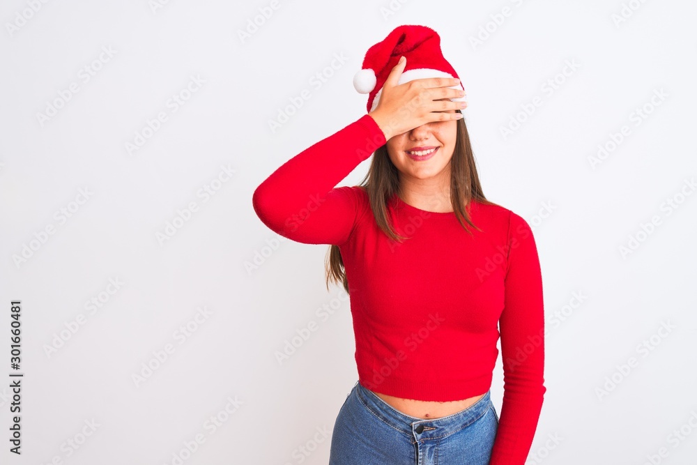 Young beautiful girl wearing Christmas Santa hat standing over isolated white background smiling and laughing with hand on face covering eyes for surprise. Blind concept. Young beautiful girl wearing Christmas Santa hat standing over isolated white background smiling and laughing with hand on face covering eyes for surprise. Blind concept.