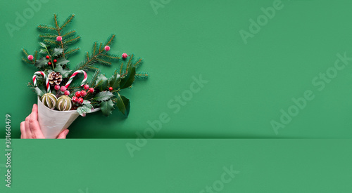 Christmas panoramic background flat lay on green paper with copy-space. Female hand holding veneer cone with fir and holly twigs with candy canes, dry fruit and berries.
