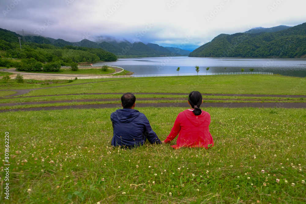Back view of Romantic Couple in love sitting on grass field and watch the lake and mountain landscape in morning. Couple love dating in holiday trip concept at Japan.