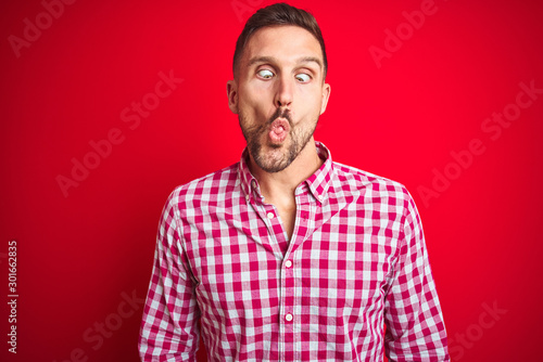 Young handsome man over red isolated background making fish face with lips, crazy and comical gesture. Funny expression.