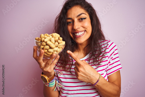 Young beautiful woman holding bowl with peanuts over isolated pink background very happy pointing with hand and finger