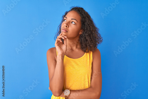 Young brazilian woman wearing yellow t-shirt standing over isolated blue background with hand on chin thinking about question, pensive expression. Smiling with thoughtful face. Doubt concept.