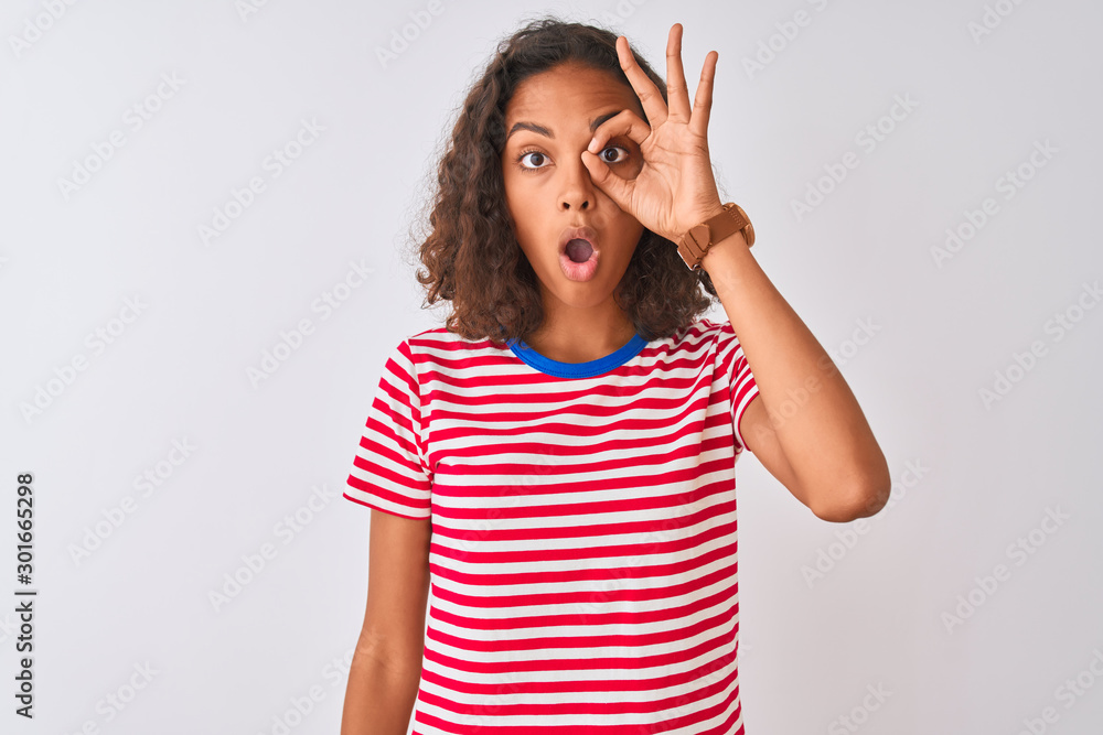 Young brazilian woman wearing red striped t-shirt standing over isolated white background doing ok gesture shocked with surprised face, eye looking through fingers. Unbelieving expression. Young brazilian woman wearing red striped t-shirt standing over isolated white background doing ok gesture shocked with surprised face, eye looking through fingers. Unbelieving expression.