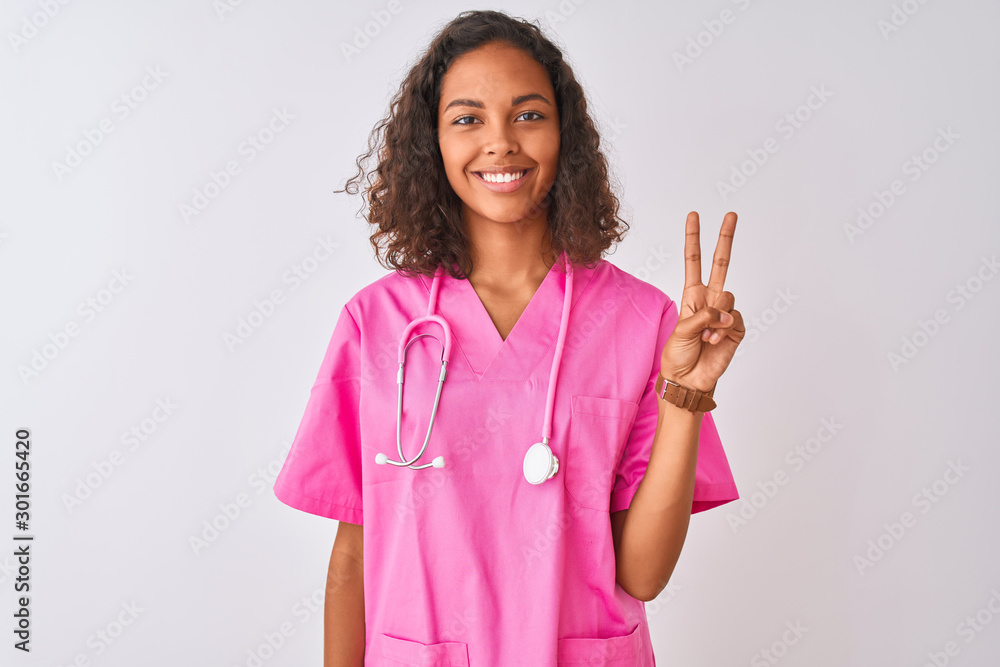 Young brazilian nurse woman wearing stethoscope standing over isolated white background showing and pointing up with fingers number two while smiling confident and happy. Young brazilian nurse woman wearing stethoscope standing over isolated white background showing and pointing up with fingers number two while smiling confident and happy.