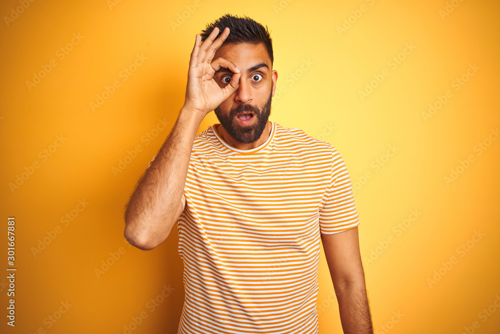 Young indian man wearing t-shirt standing over isolated yellow background doing ok gesture shocked with surprised face, eye looking through fingers. Unbelieving expression. Young indian man wearing t-shirt standing over isolated yellow background doing ok gesture shocked with surprised face, eye looking through fingers. Unbelieving expression.