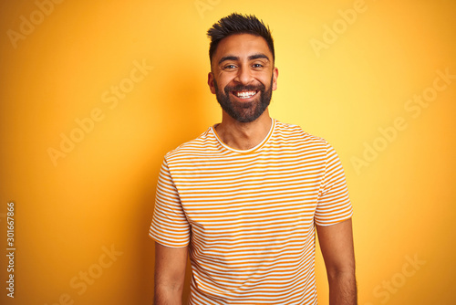 Young indian man wearing t-shirt standing over isolated yellow background with a happy and cool smile on face. Lucky person.