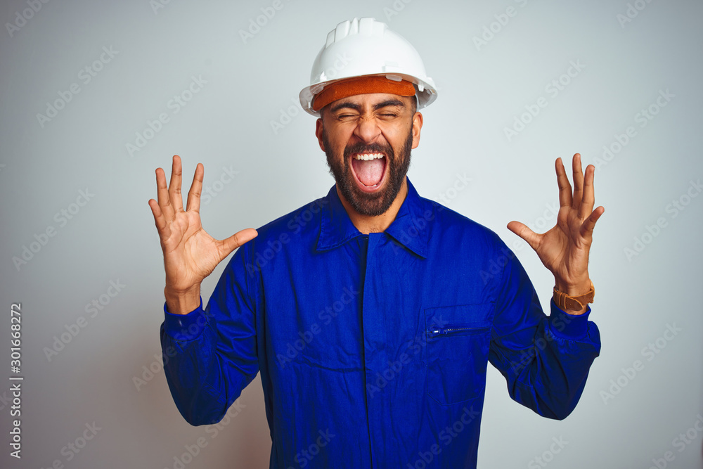 Handsome indian worker man wearing uniform and helmet over isolated white background celebrating mad and crazy for success with arms raised and closed eyes screaming excited. Winner concept