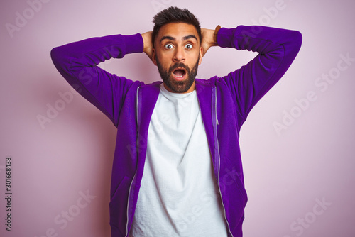Young indian man wearing purple sweatshirt standing over isolated pink background Crazy and scared with hands on head, afraid and surprised of shock with open mouth