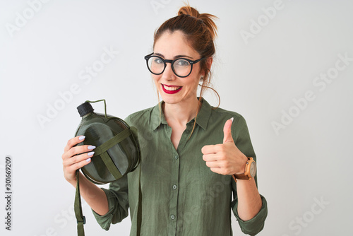 Beautiful redhead woman wearing glasses holding canteen over isolated white background happy with big smile doing ok sign, thumb up with fingers, excellent sign