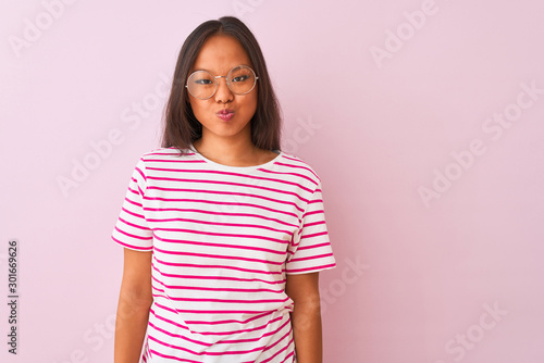Young chinese woman wearing striped t-shirt and glasses over isolated pink background puffing cheeks with funny face. Mouth inflated with air, crazy expression.