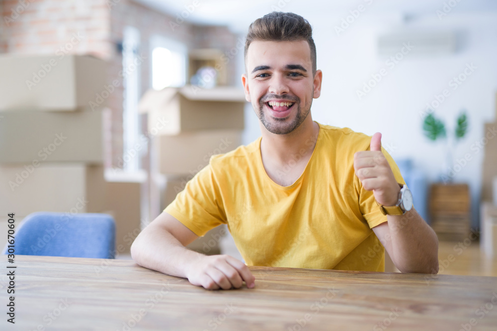 Fototapeta premium Young man sitting on the table with cardboard boxes behind him moving to new home doing happy thumbs up gesture with hand. Approving expression looking at the camera with showing success.