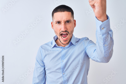 Young handsome business man standing over isolated background angry and mad raising fist frustrated and furious while shouting with anger. Rage and aggressive concept.