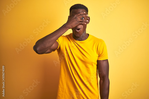 Young african american man wearing casual t-shirt standing over isolated yellow background peeking in shock covering face and eyes with hand, looking through fingers with embarrassed expression.