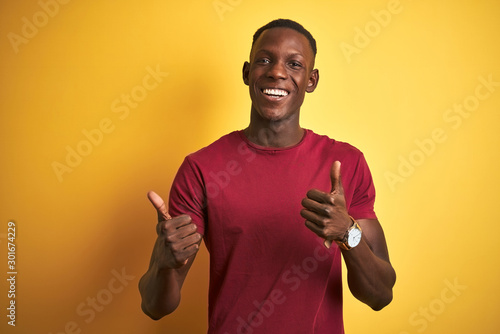 Young african american man wearing red t-shirt standing over isolated yellow background success sign doing positive gesture with hand, thumbs up smiling and happy. Cheerful expression and winner