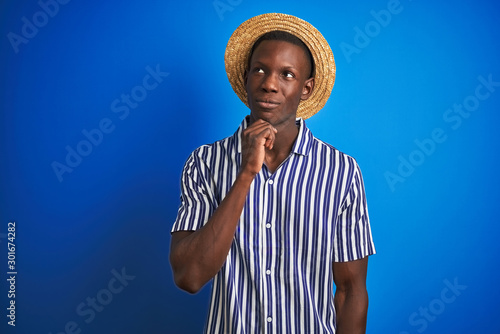 African american man wearing striped shirt and summer hat over isolated blue background with hand on chin thinking about question, pensive expression. Smiling with thoughtful face. Doubt concept.