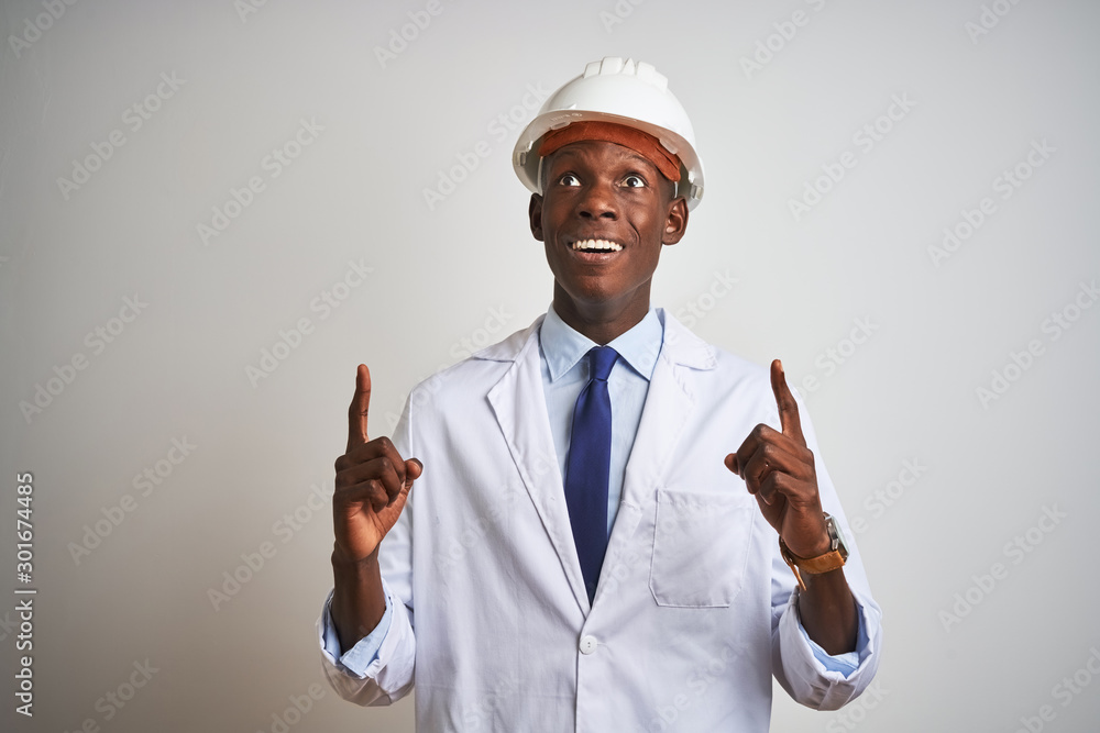 African american engineer man wearing coat and helmet over isolated white background amazed and surprised looking up and pointing with fingers and raised arms. African american engineer man wearing coat and helmet over isolated white background amazed and surprised looking up and pointing with fingers and raised arms.