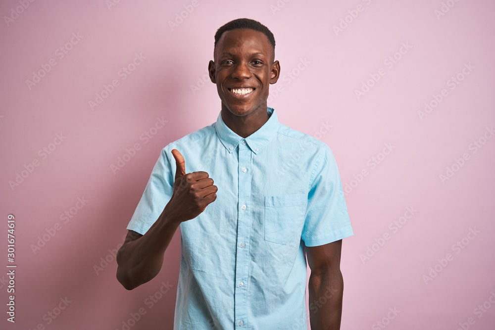 African american man wearing blue casual shirt standing over isolated pink background doing happy thumbs up gesture with hand. Approving expression looking at the camera showing success. African american man wearing blue casual shirt standing over isolated pink background doing happy thumbs up gesture with hand. Approving expression looking at the camera showing success.