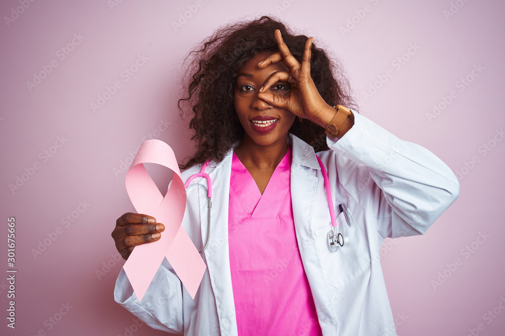 Young african american doctor woman holding cancer ribbon over isolated pink background with happy face smiling doing ok sign with hand on eye looking through fingers Young african american doctor woman holding cancer ribbon over isolated pink background with happy face smiling doing ok sign with hand on eye looking through fingers