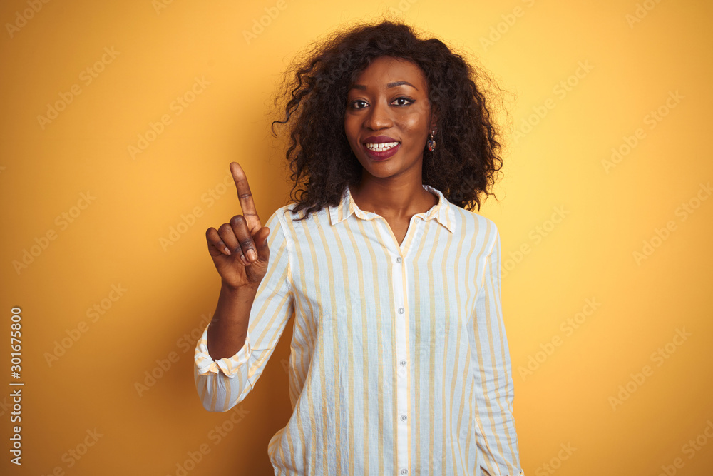 African american woman wearing striped shirt standing over isolated yellow background showing and pointing up with finger number one while smiling confident and happy. African american woman wearing striped shirt standing over isolated yellow background showing and pointing up with finger number one while smiling confident and happy.