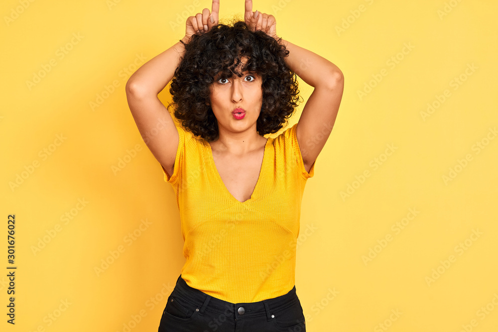 Young arab woman with curly hair wearing t-shirt standing over isolated yellow background doing funny gesture with finger over head as bull horns Young arab woman with curly hair wearing t-shirt standing over isolated yellow background doing funny gesture with finger over head as bull horns