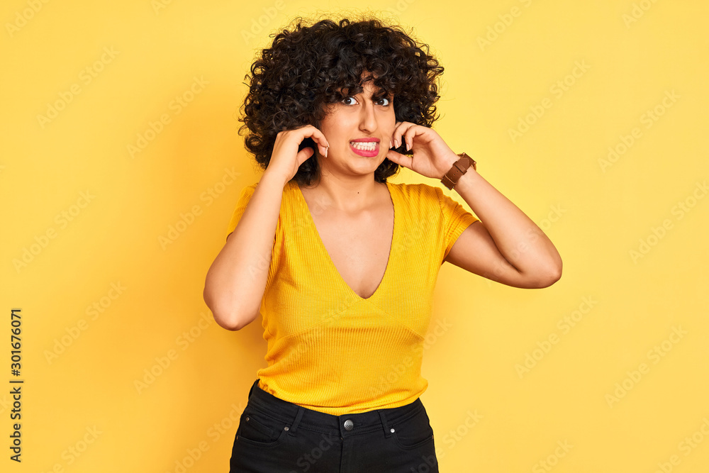 Young arab woman with curly hair wearing t-shirt standing over isolated yellow background covering ears with fingers with annoyed expression for the noise of loud music. Deaf concept.