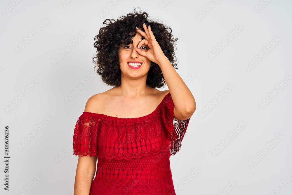 Young arab woman with curly hair wearing red dress standing over isolated white background doing ok gesture with hand smiling, eye looking through fingers with happy face. Young arab woman with curly hair wearing red dress standing over isolated white background doing ok gesture with hand smiling, eye looking through fingers with happy face.