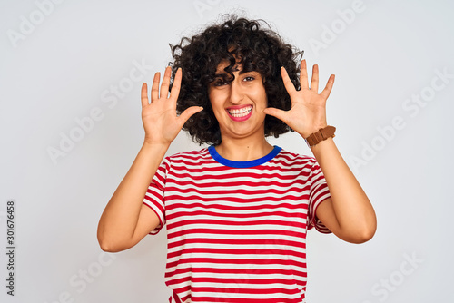 Young arab woman with curly hair wearing striped t-shirt over isolated white background showing and pointing up with fingers number ten while smiling confident and happy.