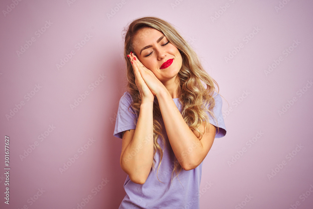 Young beautiful woman wearing purple t-shirt standing over pink isolated background sleeping tired dreaming and posing with hands together while smiling with closed eyes. Young beautiful woman wearing purple t-shirt standing over pink isolated background sleeping tired dreaming and posing with hands together while smiling with closed eyes.