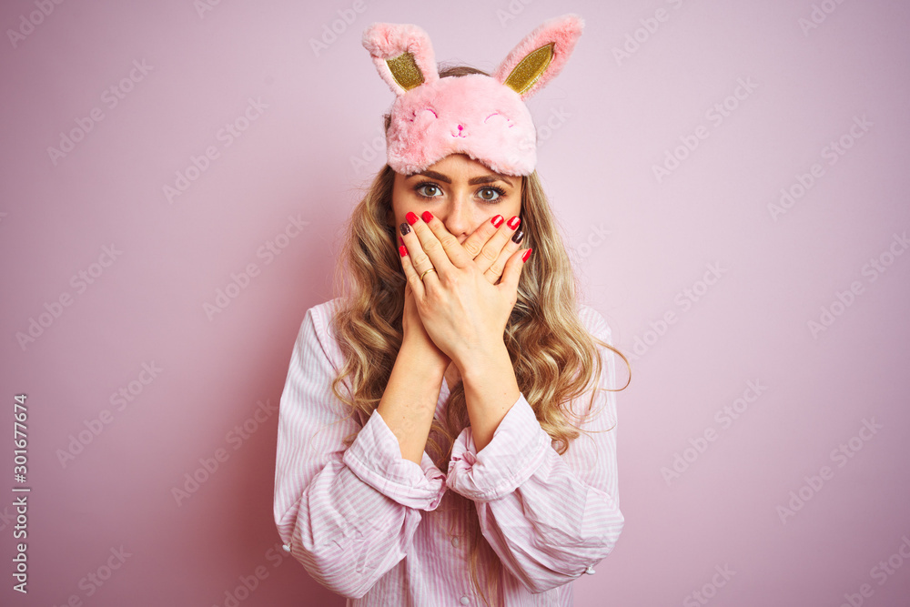 Young beautiful woman wearing pajama and sleep mask over pink isolated background shocked covering mouth with hands for mistake. Secret concept. Young beautiful woman wearing pajama and sleep mask over pink isolated background shocked covering mouth with hands for mistake. Secret concept.